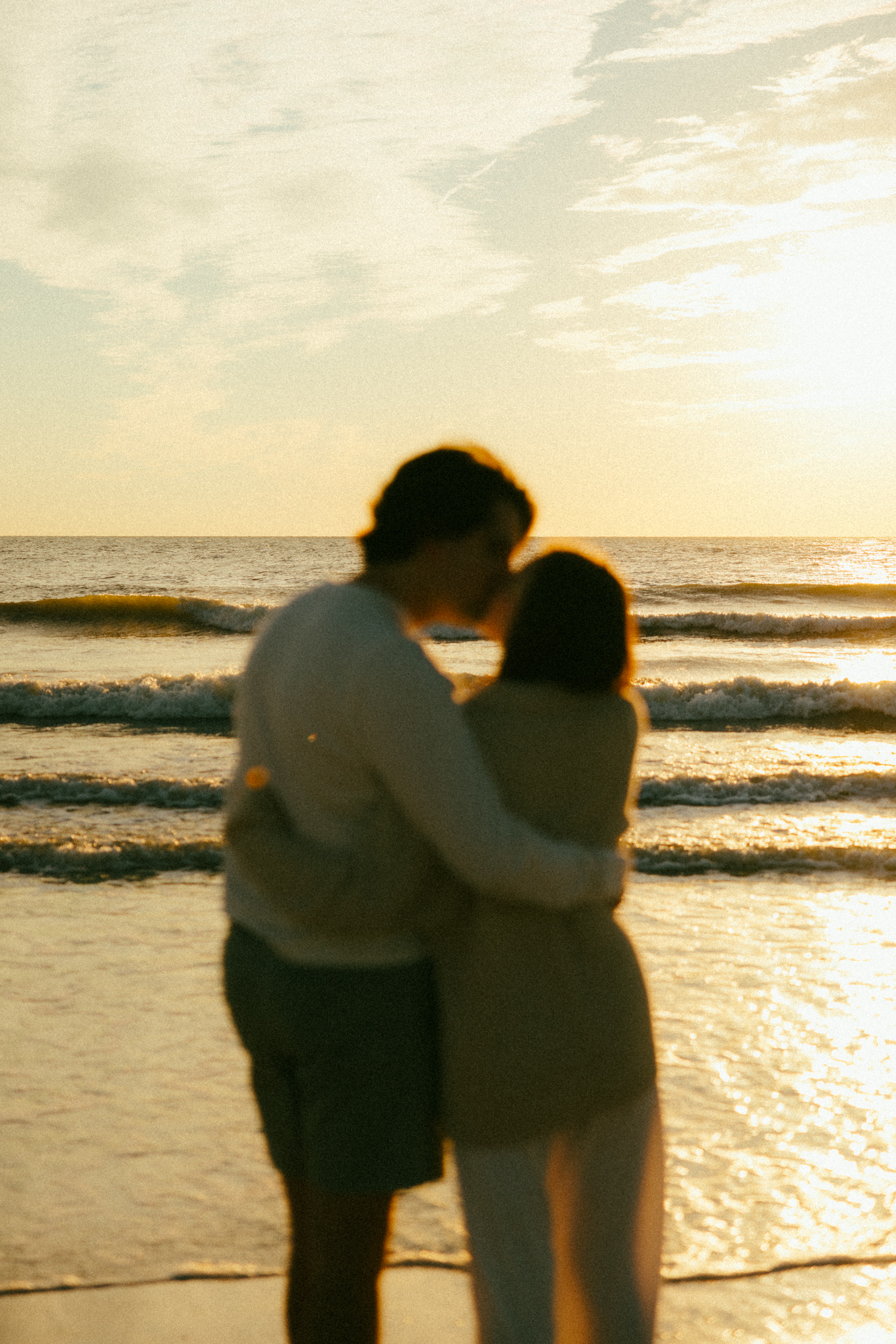 Documentary style photo of couple kissing with the camera focused on the waves behind them at sunset.