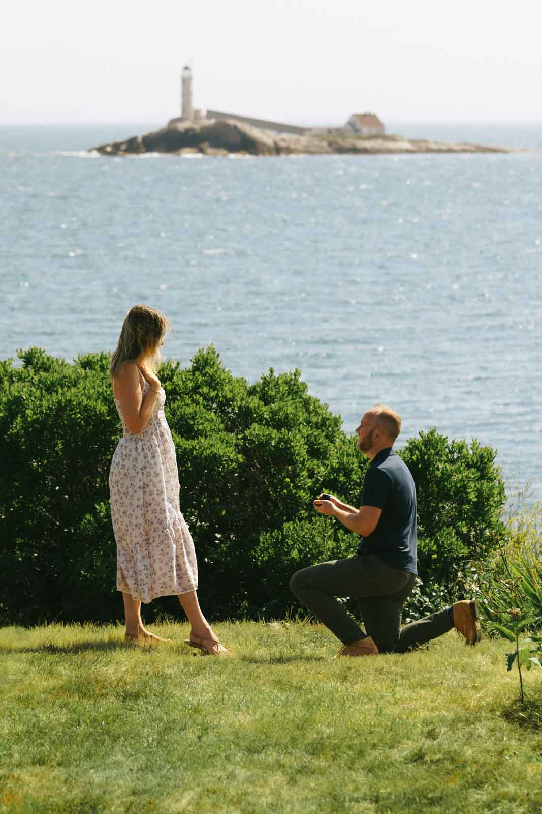 Man proposing to his girlfriend on Star Island, NH. The ocean and a lighthouse is in the background.