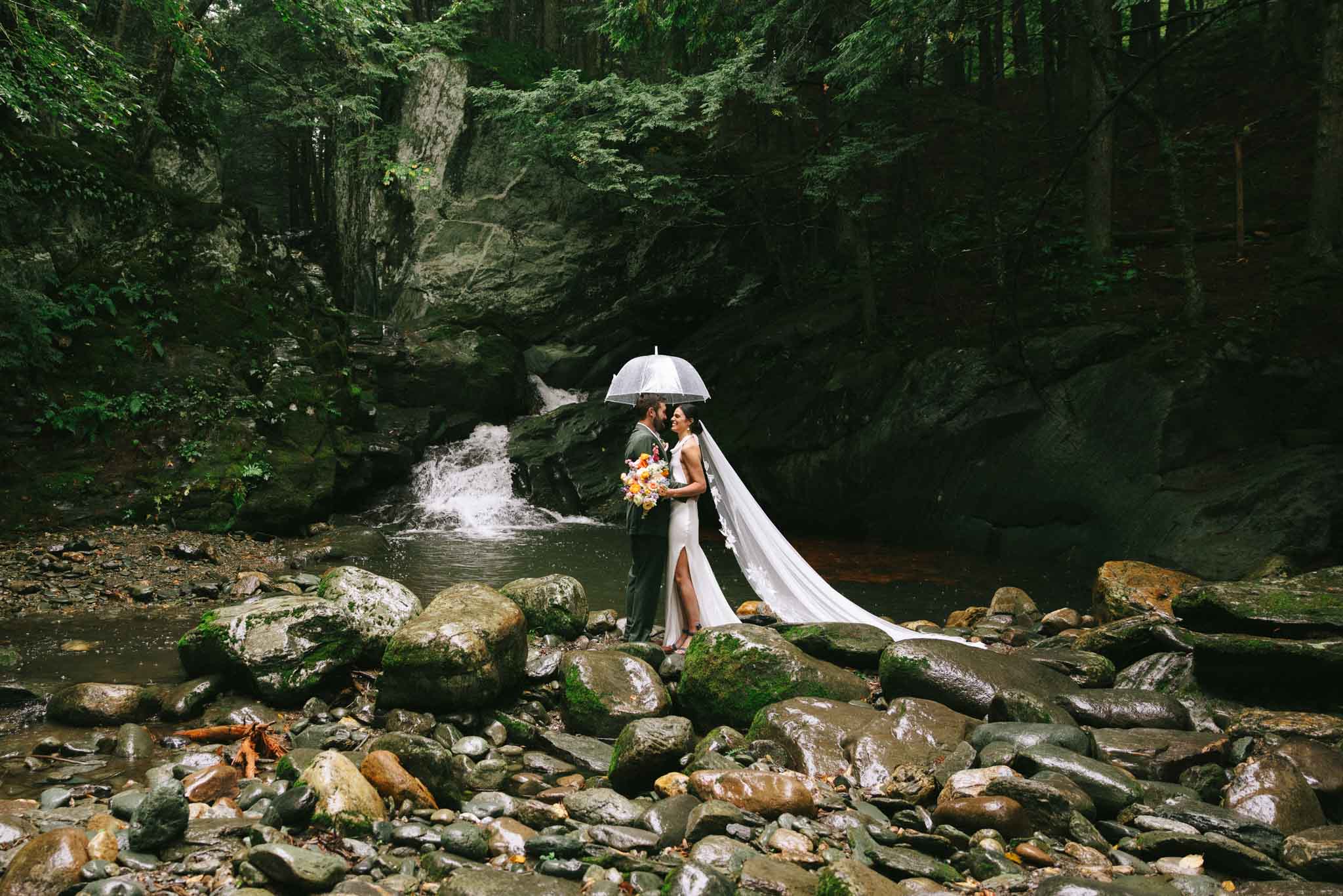 Bride and groom stand with an umbrella in front of Madbush Falls in Waitsfield, VT.
