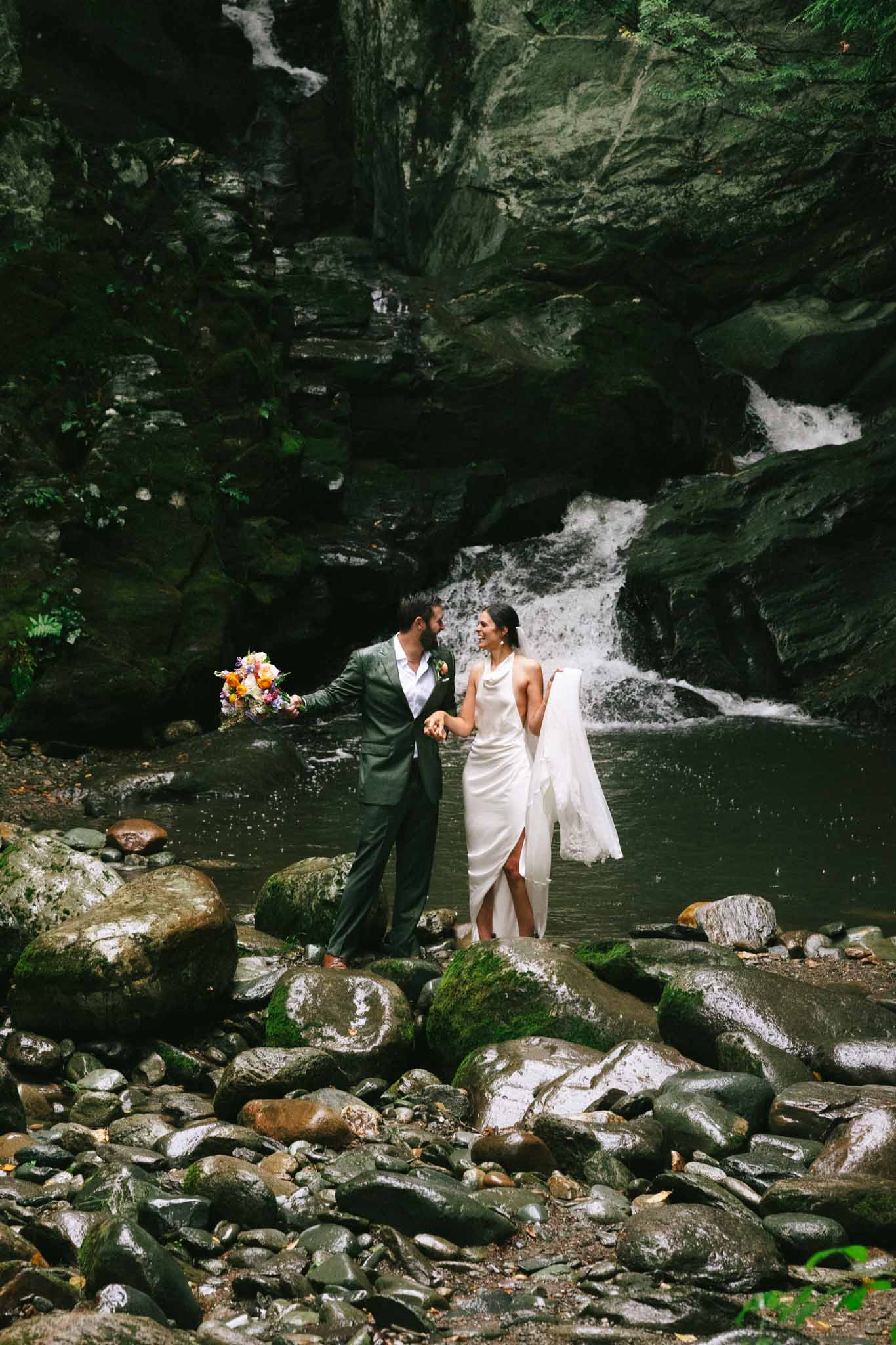 Bride and groom stand in front of Madbush Falls in Vermont